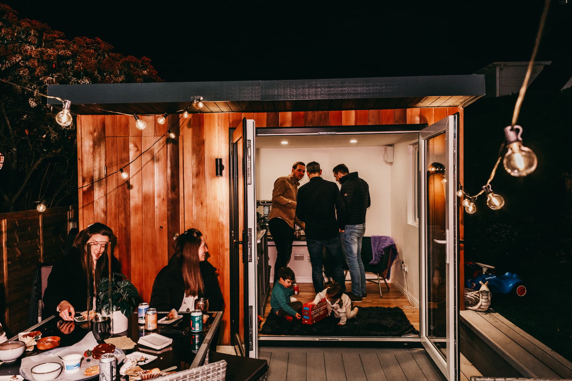 Cedar garden room lit up at night with family gathered inside and friends dining on the deck