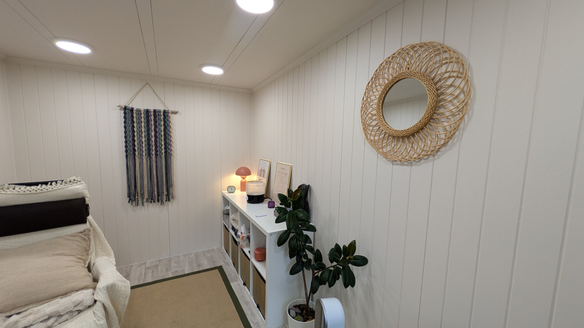 Calm white panelled therapy room in a garden building with rattan mirror and treatment couch