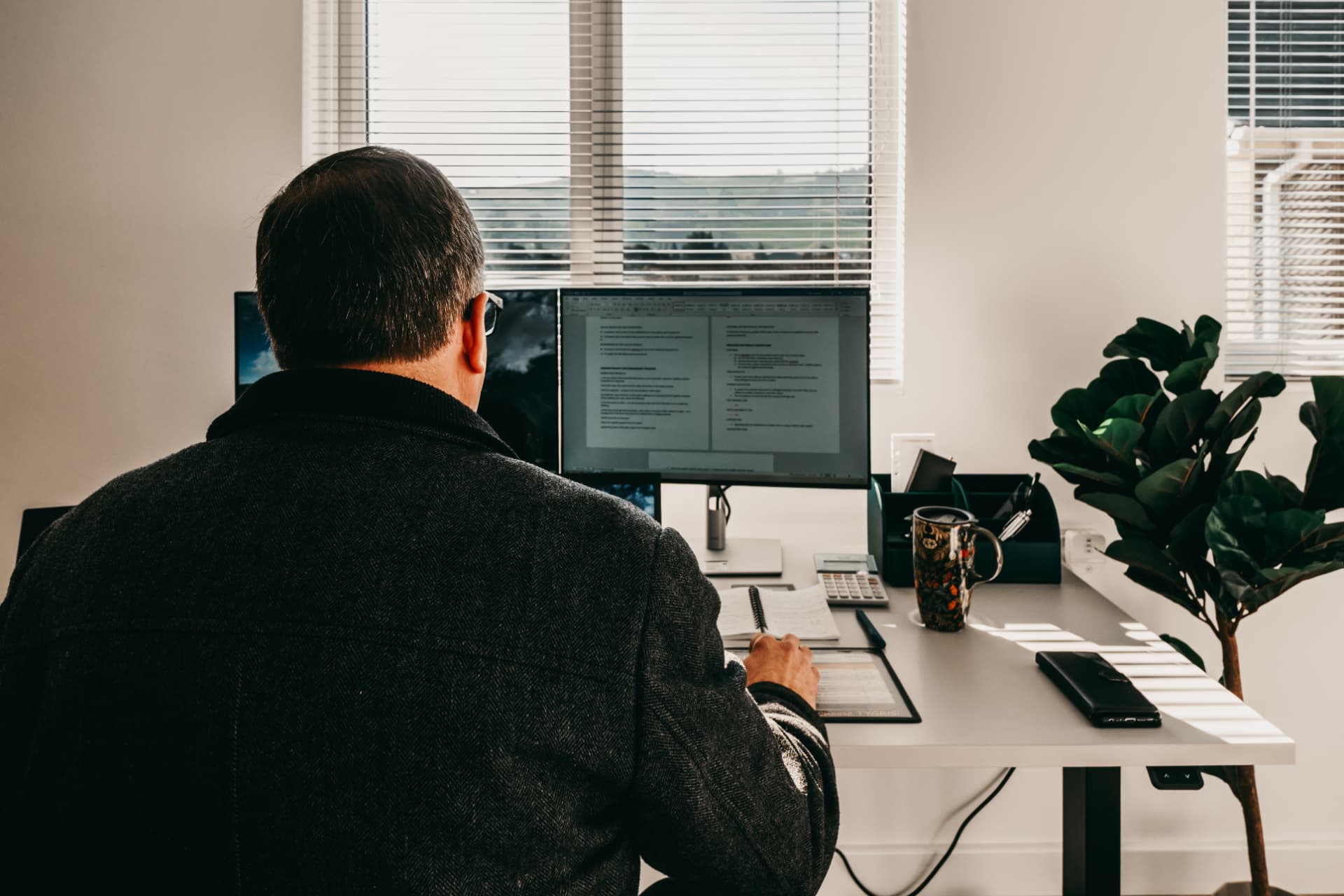 Man working at a dual-monitor desk in a bright garden office with window blinds