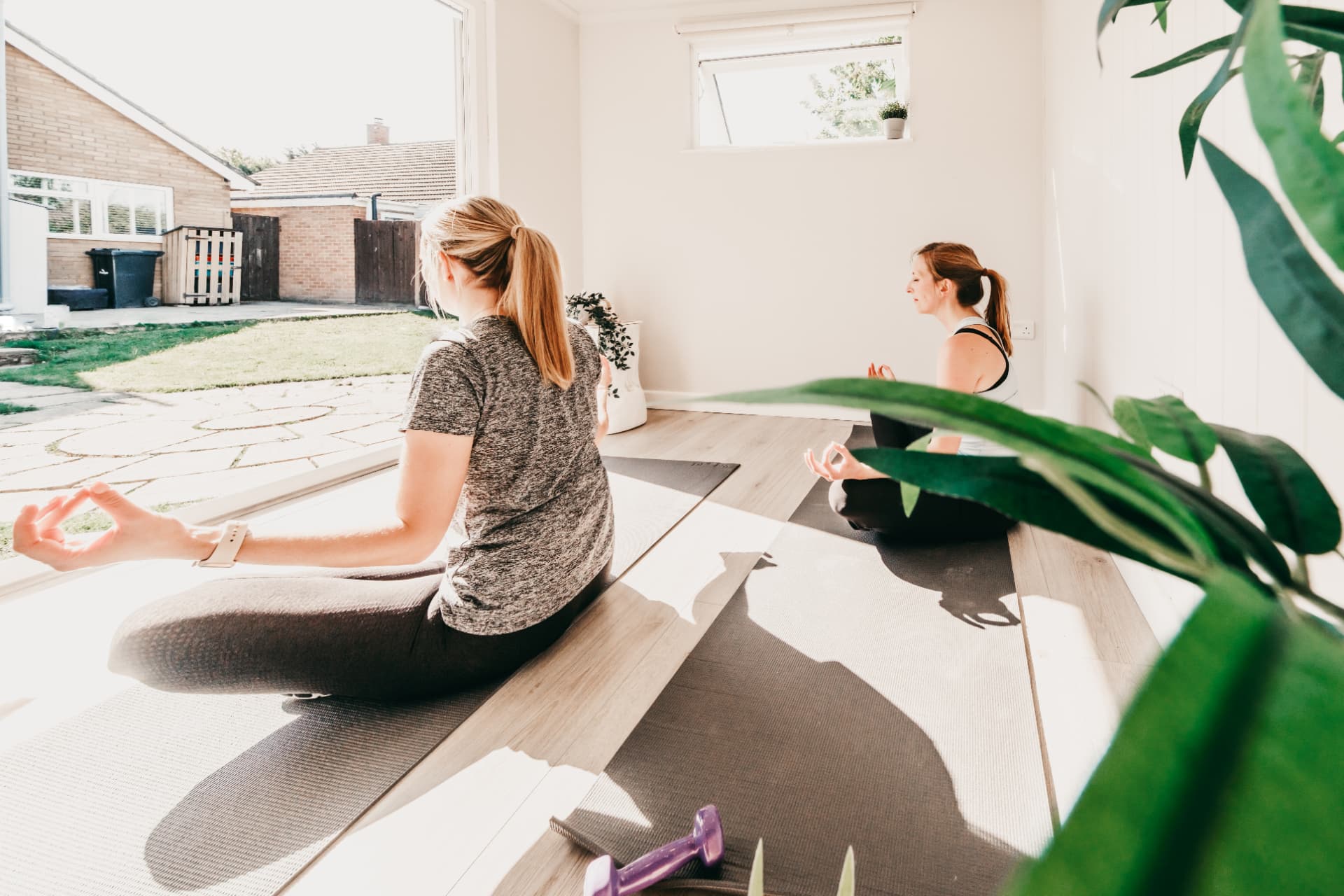 Two women meditating on mats in a bright garden yoga studio with doors open