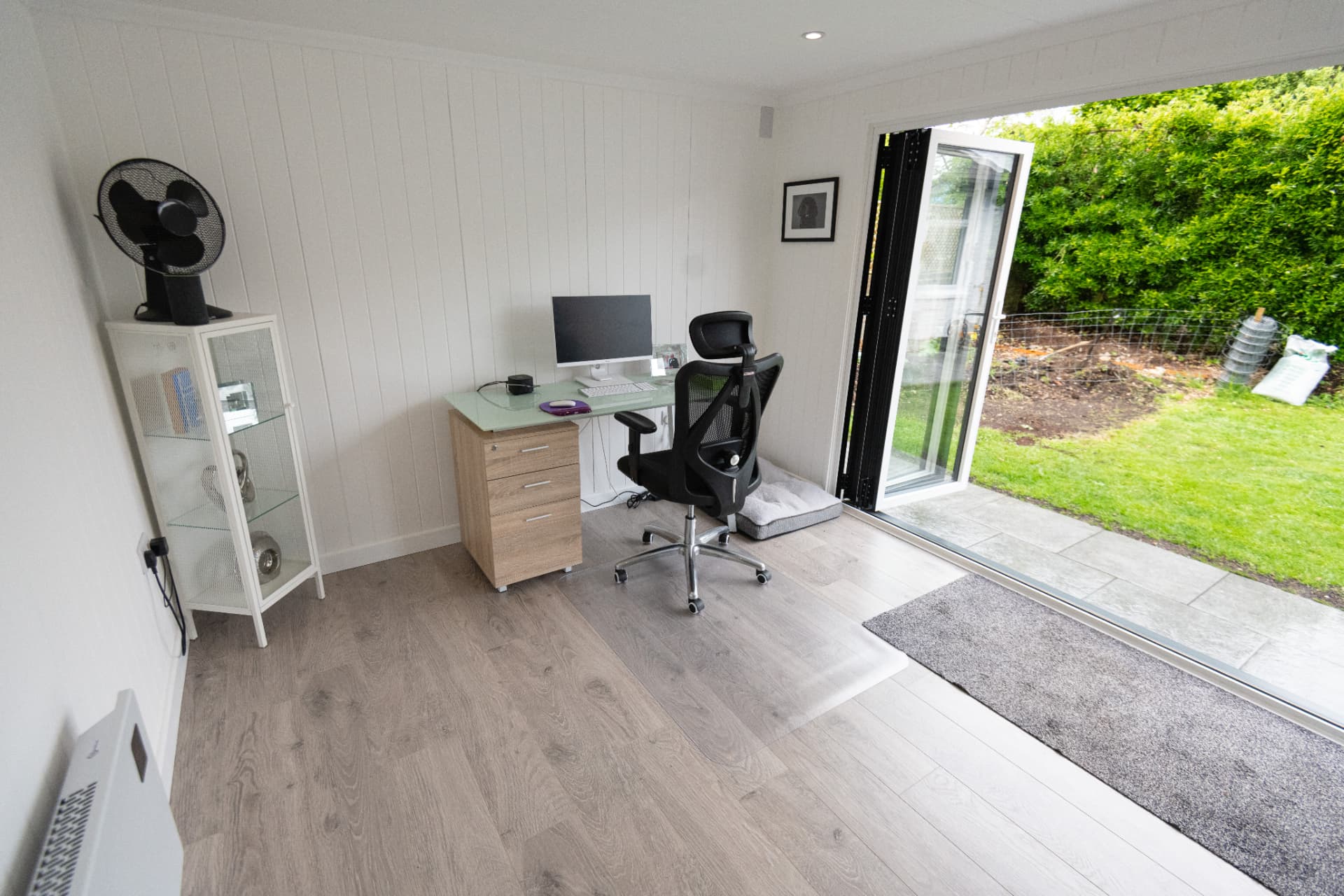 White panelled garden office with a glass desk and bifold doors open to the lawn