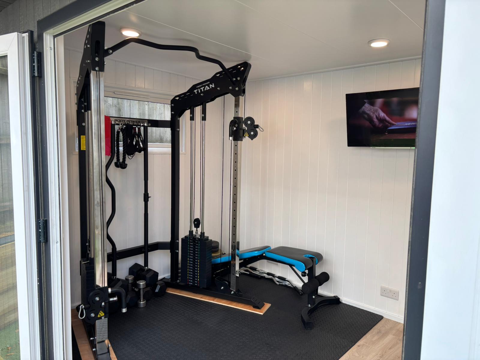 Garden gym with a Titan multi-gym, weight bench and wall-mounted TV seen through open doors