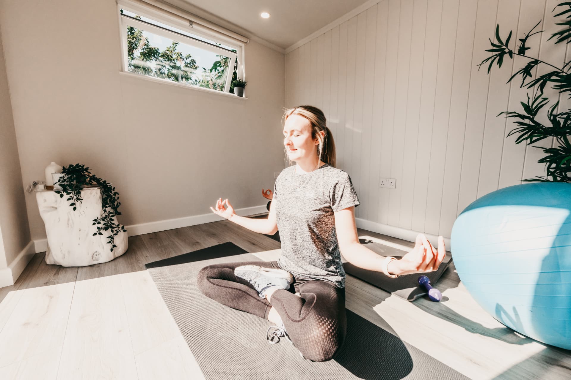 Woman meditating cross-legged on a mat in a sunlit garden yoga room