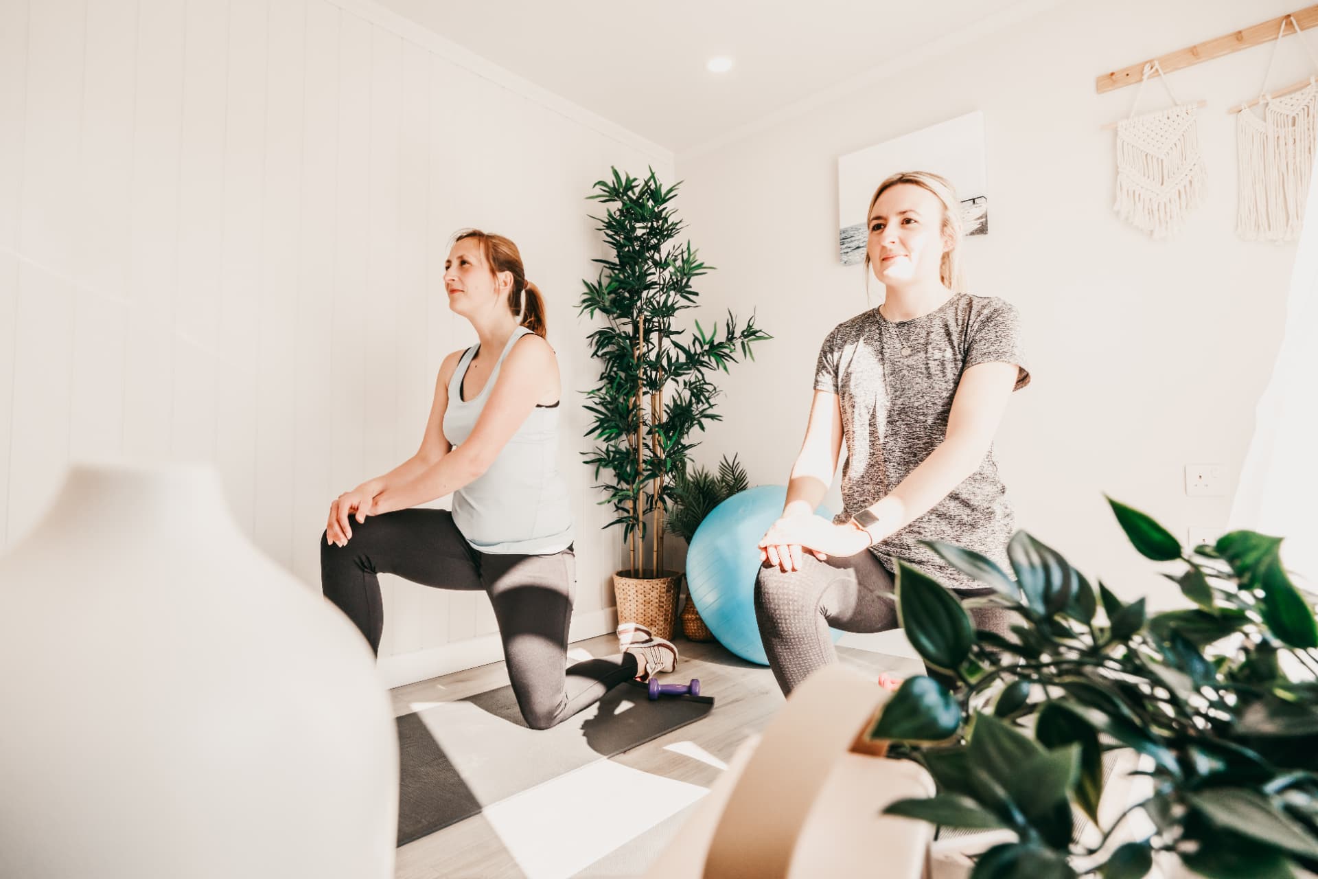 Two women stretching in a garden yoga room with plants and macrame wall hangings