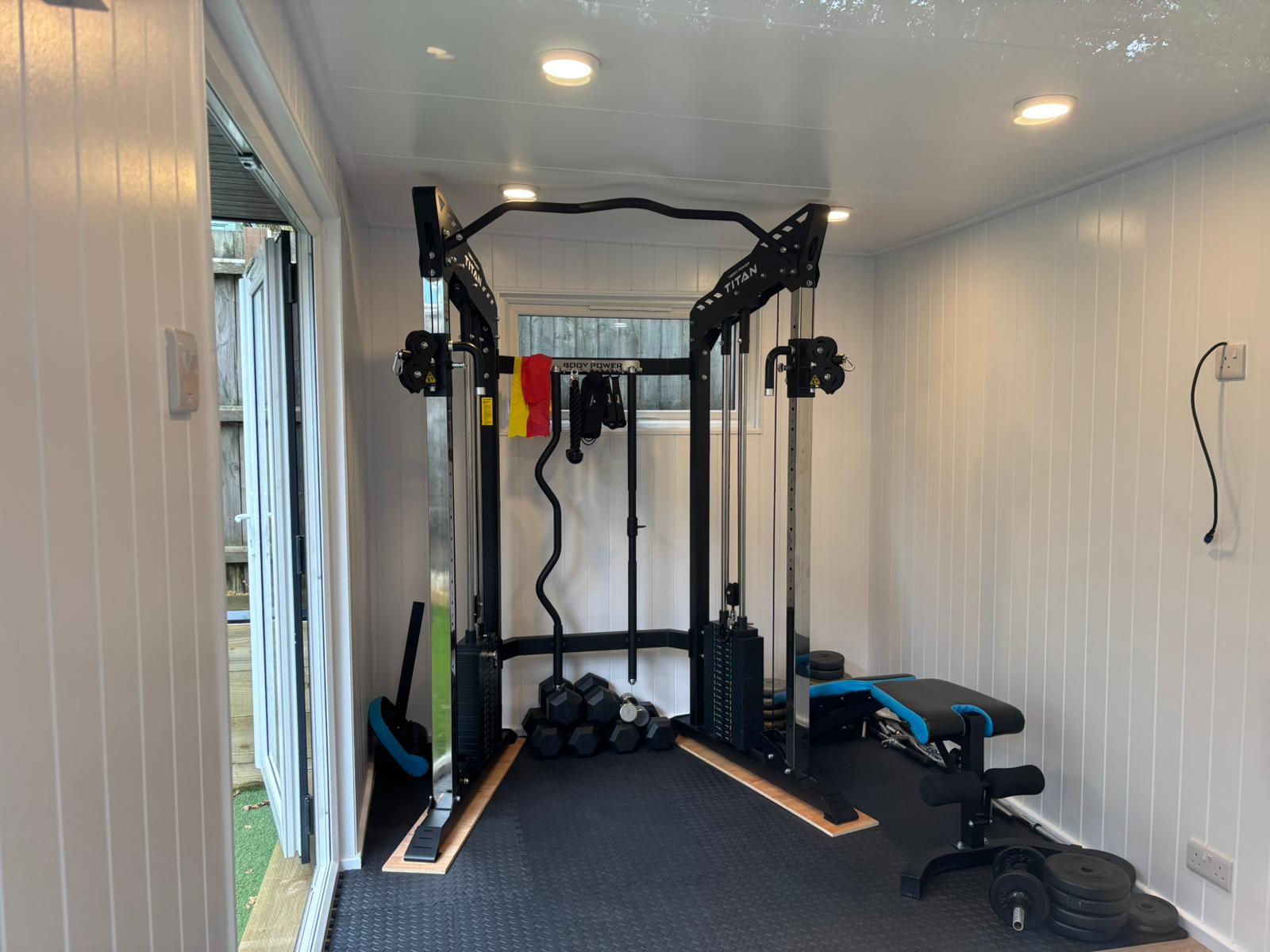 Garden gym interior with a Titan cable machine, dumbbells and adjustable bench